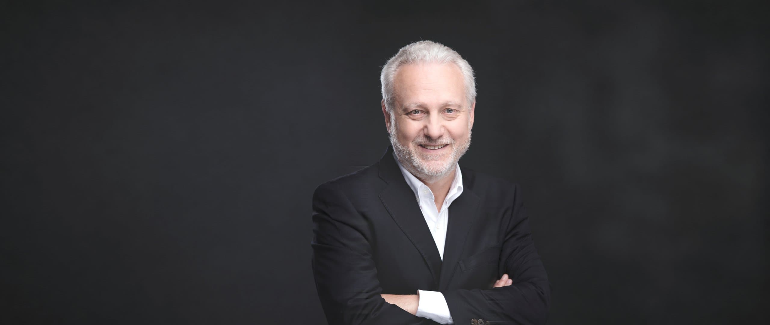 Yves Bigot smiling with arms crossed, in a dark jacket and white shirt against a dark background